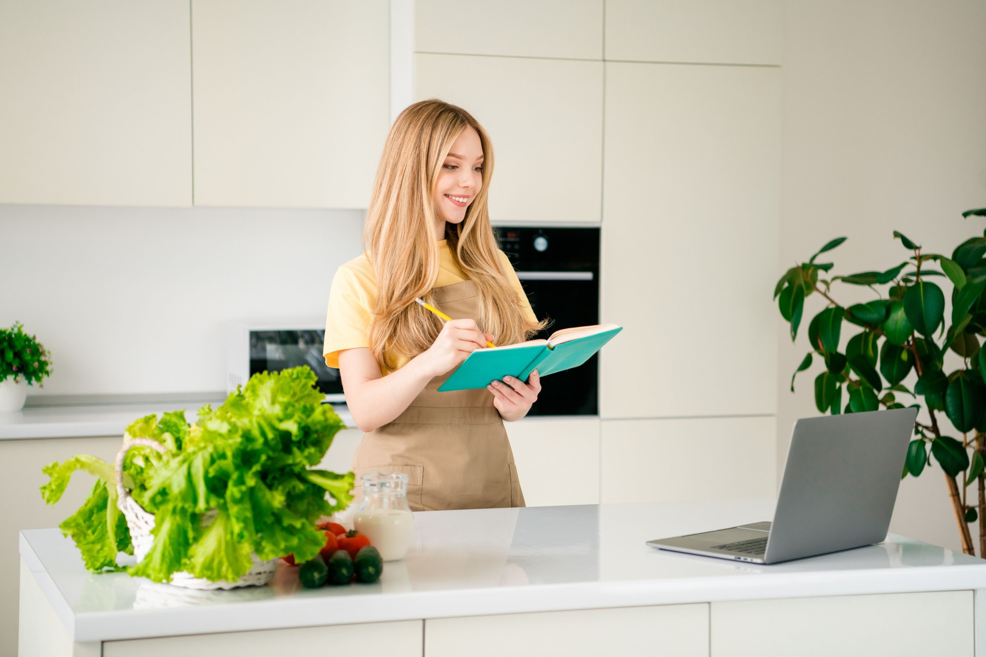 Photo of nice teen girl write recipe notebook laptop dressed apron making domestic homemade dinner interior kitchen house apartment indoors